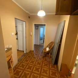 Michał Rostowski - Korytarz w mieszkaniu z brązowymi ścianami i podłogą w kwadraty, z widoczną starą, opartą o ścianę szarą płytą drzwiową i wiklinowym fotelem.