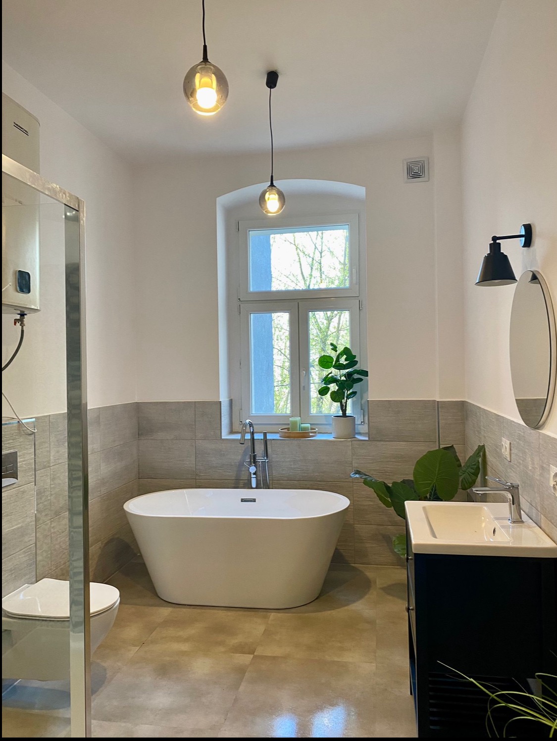 Jasna łazienka po remoncie z wolnostojącą wanną, umywalką z szafką w kolorze granatowym, szarą glazurą do połowy ściany, okrągłym lustrem i dwiema wiszącymi lampami kulami.