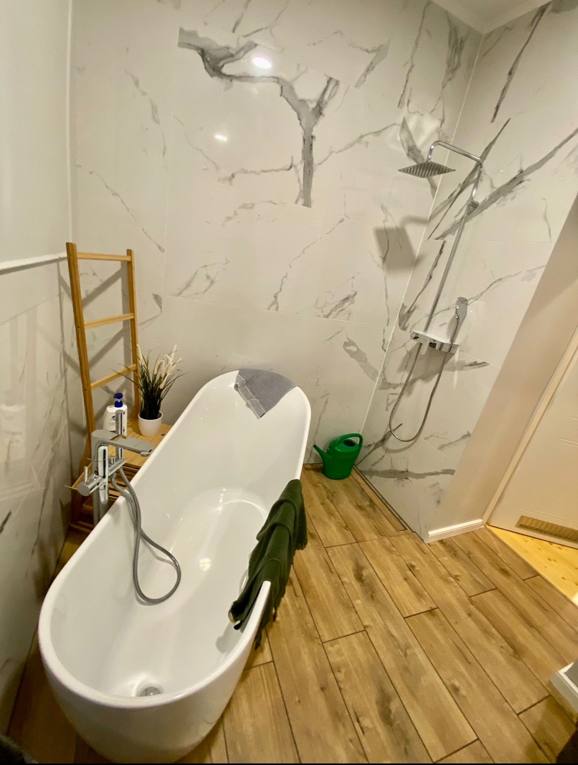 Nowoczesna łazienka z wolnostojącą białą wanną, marmurowymi płytkami na ścianach i drewnopodobną podłogą. Widoczny ręcznik na wannie, drabinka dekoracyjna i zielona konewka.