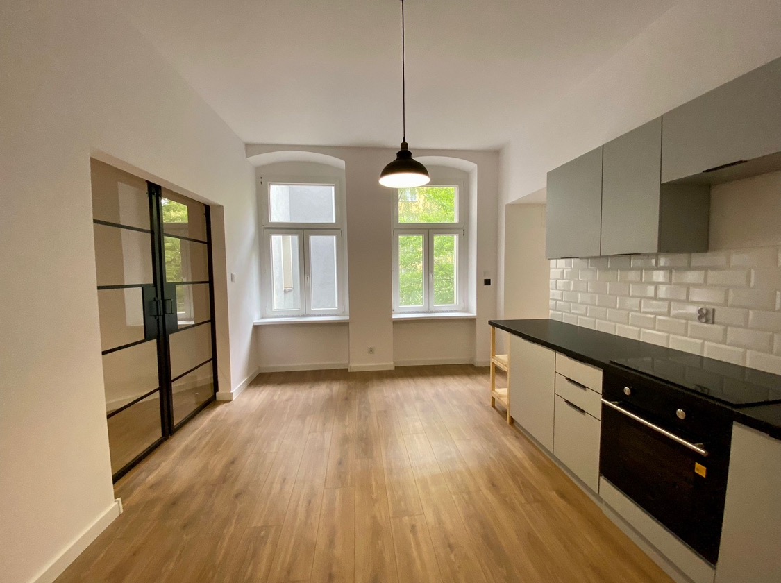 Wykończona kuchnia w jasnych barwach z drewnianą podłogą, szarymi szafkami i czarnym blatem, widok na okno z zielenią za oknem oraz czarne, przeszklone drzwi przesuwne.