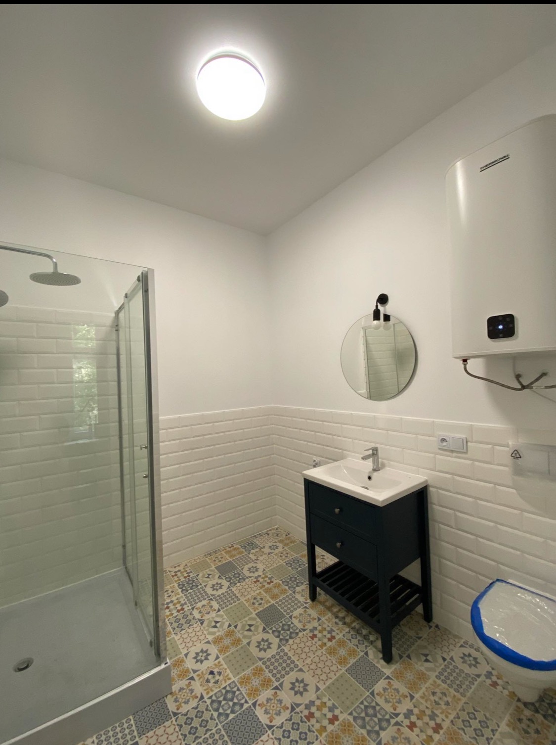 Nowoczesna łazienka po remoncie z kabiną prysznicową, umywalką z szafką w kolorze granatowym, okrągłym lustrem z oświetleniem, bojlerem i wzorzystą podłogą z płytek.