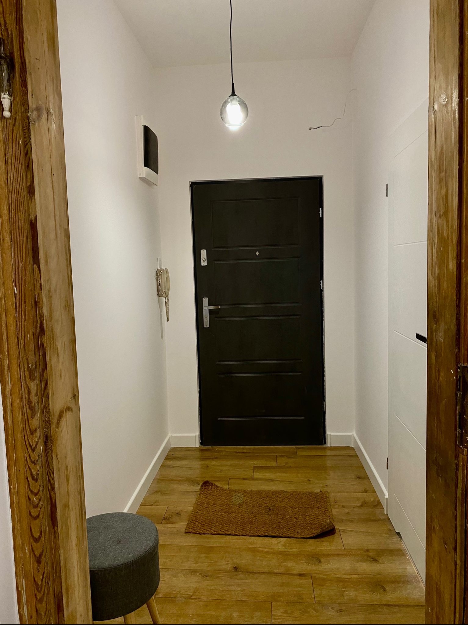 Wnętrze przedpokoju po remoncie: ciemne drzwi wejściowe, panele podłogowe imitujące drewno, szary puf, fragment drewnianej kolumny oraz białe ściany z widocznym domofonem i staromodnym telefonem.
