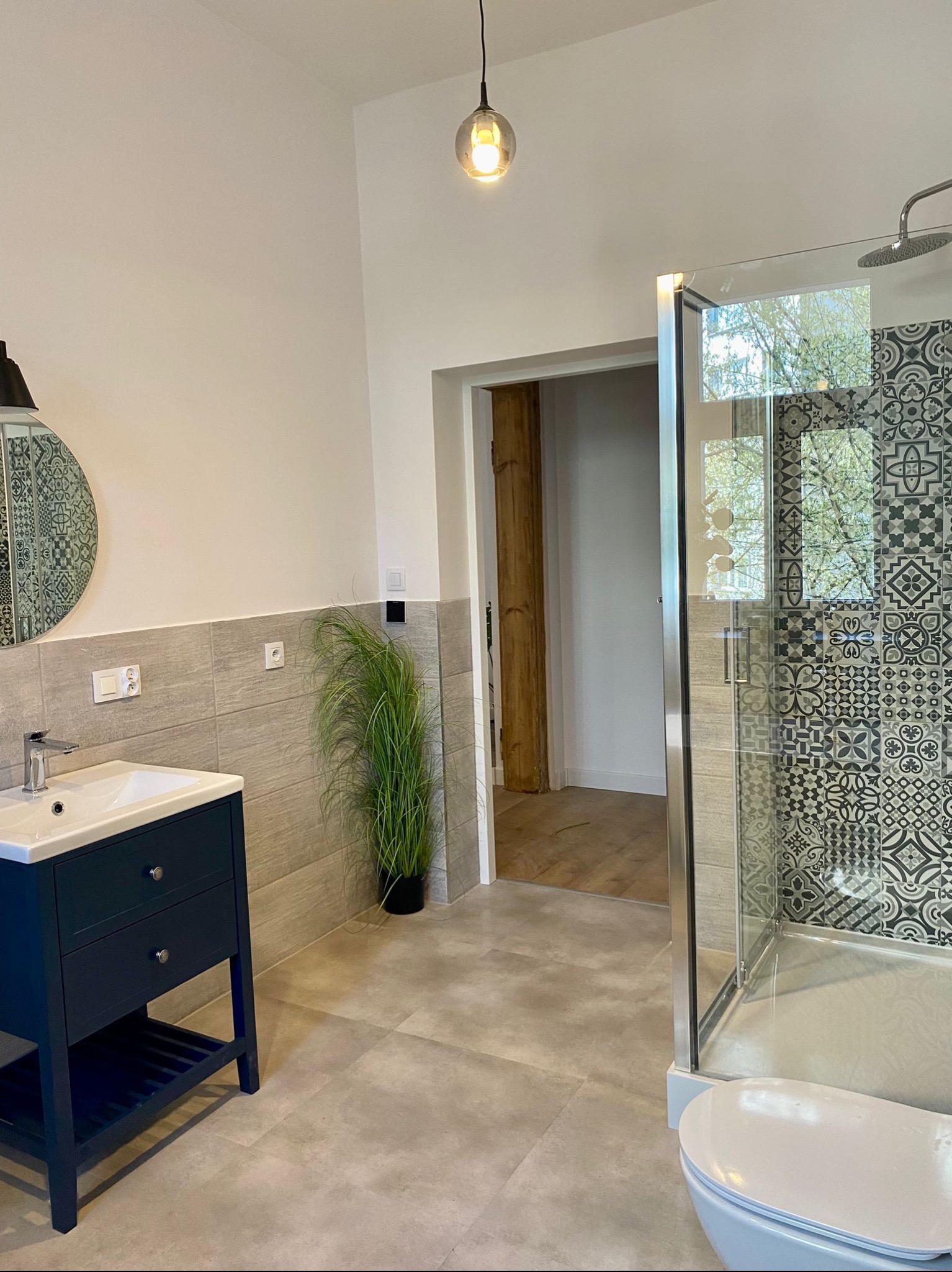 Wykończona łazienka w Gliwicach z kabiną prysznicową z wzorzystymi płytkami, umywalką z szafką w kolorze granatowym i dekoracyjną rośliną w tle.