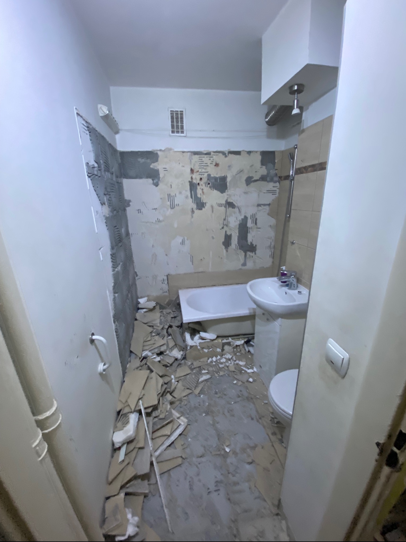 Wnętrze łazienki w trakcie demontażu: fragmenty płytek, gruzu i starej zabudowy leżą na podłodze, ściany częściowo obdarte z okładzin, widoczna wanna, umywalka i toaleta.