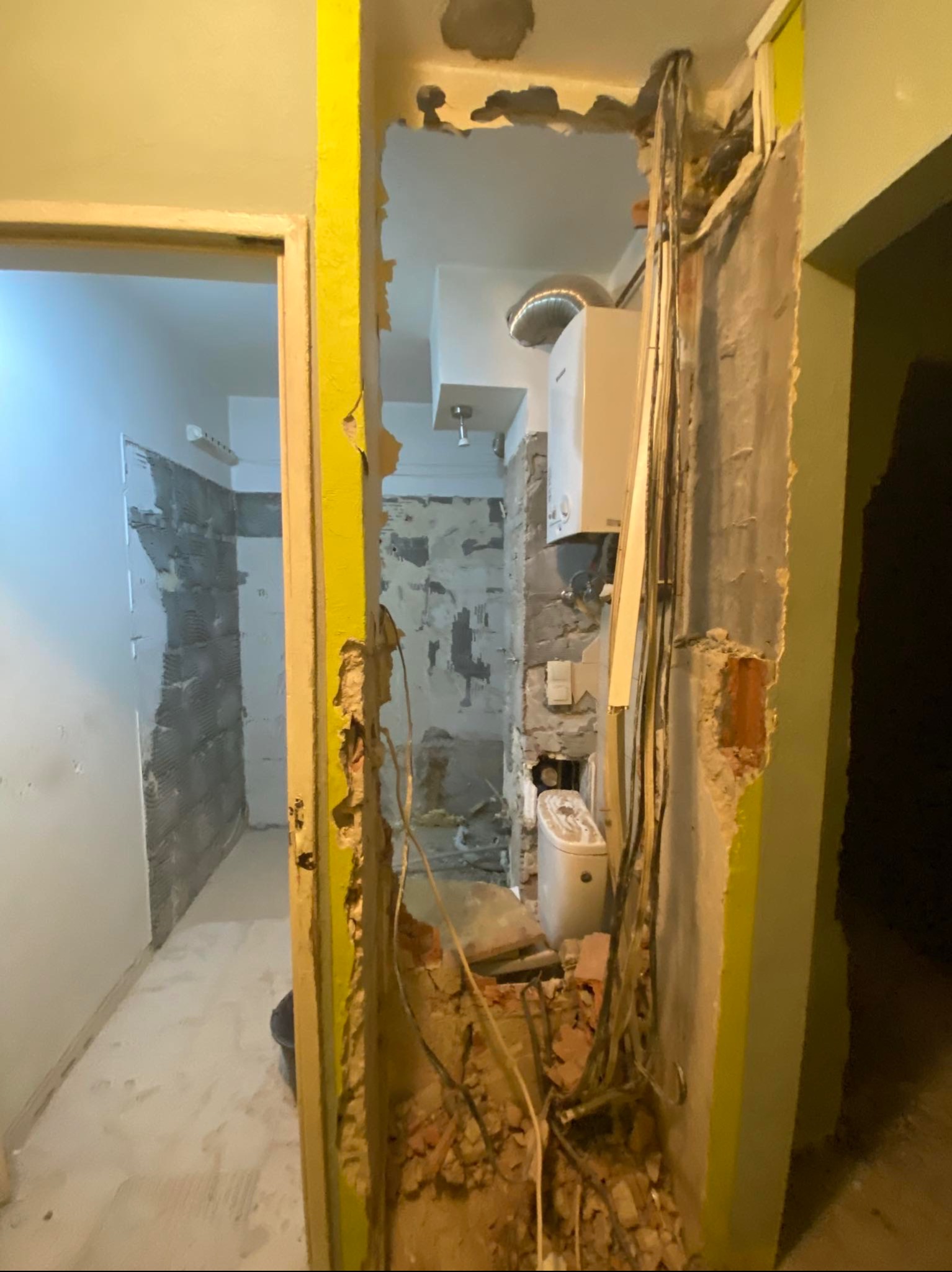 Wnętrze w trakcie demontażu ścianki działowej, odsłonięte przewody elektryczne i instalacja wodna, widoczna łazienka z częściowo położonymi płytkami i bojlerem.