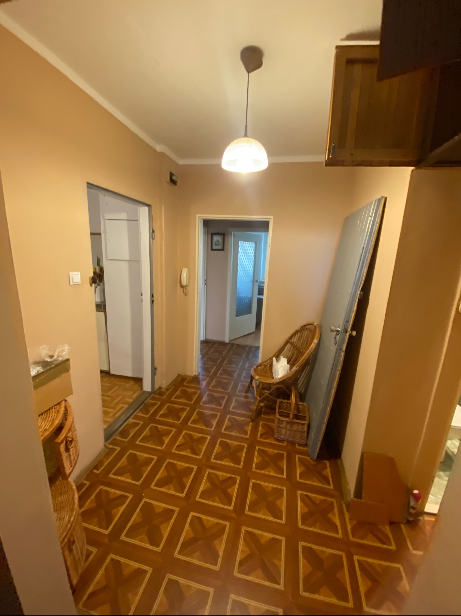 Korytarz w mieszkaniu z brązowymi ścianami i podłogą w kwadraty, z widoczną starą, opartą o ścianę szarą płytą drzwiową i wiklinowym fotelem.