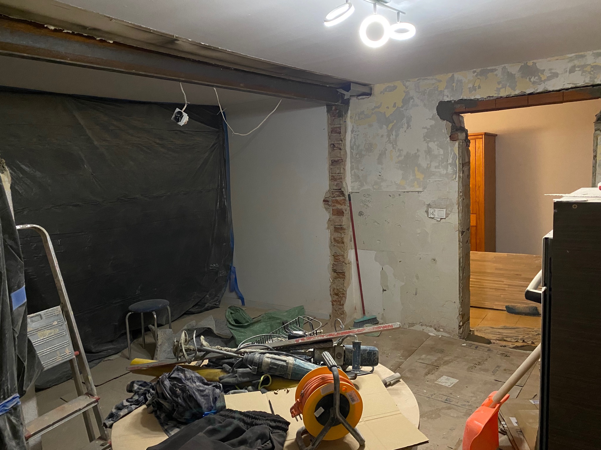 Demontaż ściany działowej w mieszkaniu, widoczny fragment ceglanej konstrukcji, wystające przewody elektryczne, zabezpieczona folią przestrzeń, narzędzia i drabina w pomieszczeniu w trakcie prac.