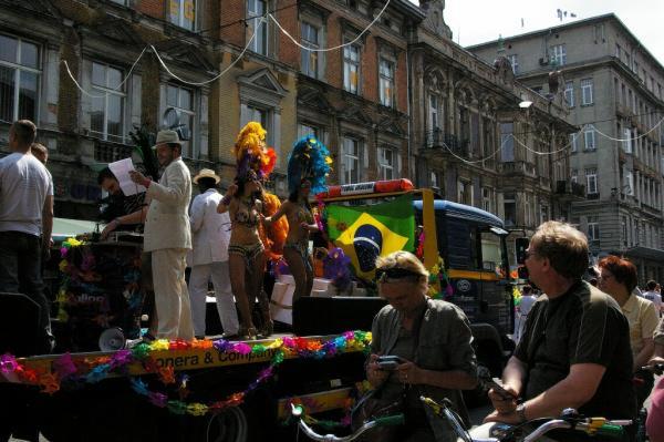 Kolorowy korowód z tancerkami w pióropuszach i flagą Brazylii na platformie ciężarówki, DJ gra muzykę, widzowie obserwują z rowerów, kamienice w tle.