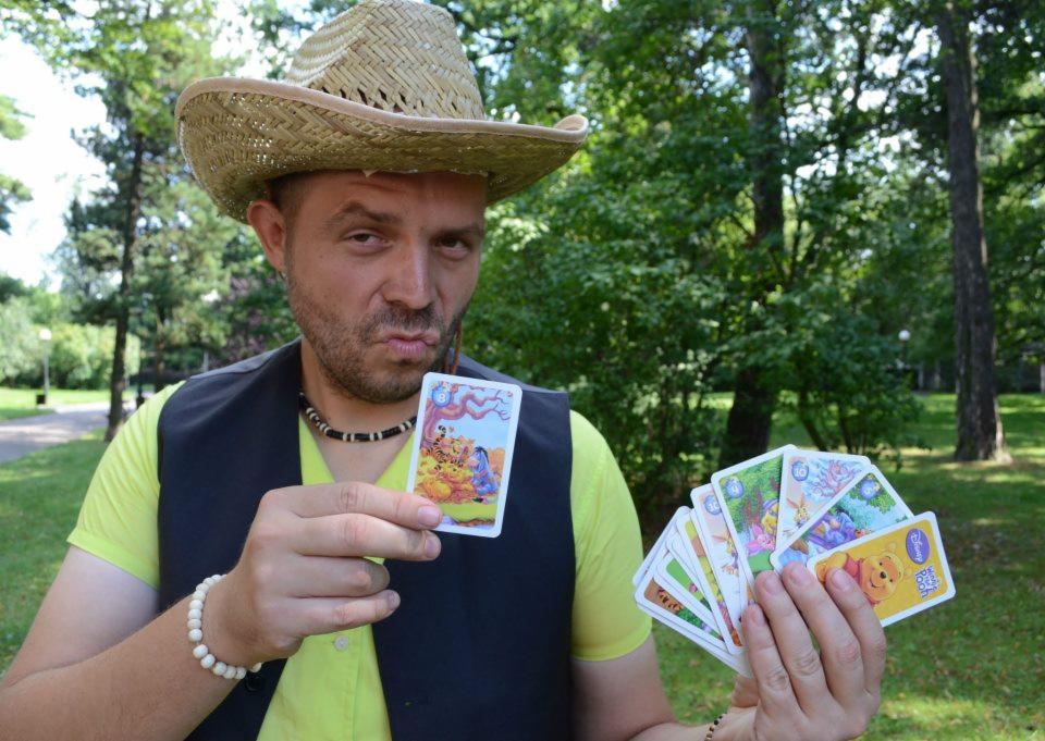 Mężczyzna w słomkowym kapeluszu prezentuje karty z postaciami z bajki Kubuś Puchatek na tle zieleni parkowej.