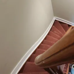 Spiralne schody z bordową wykładziną dywanową i drewnianą balustradą, widok z góry. Ściany pomalowane na jasny odcień beżu z białymi listwami przypodłogowymi.