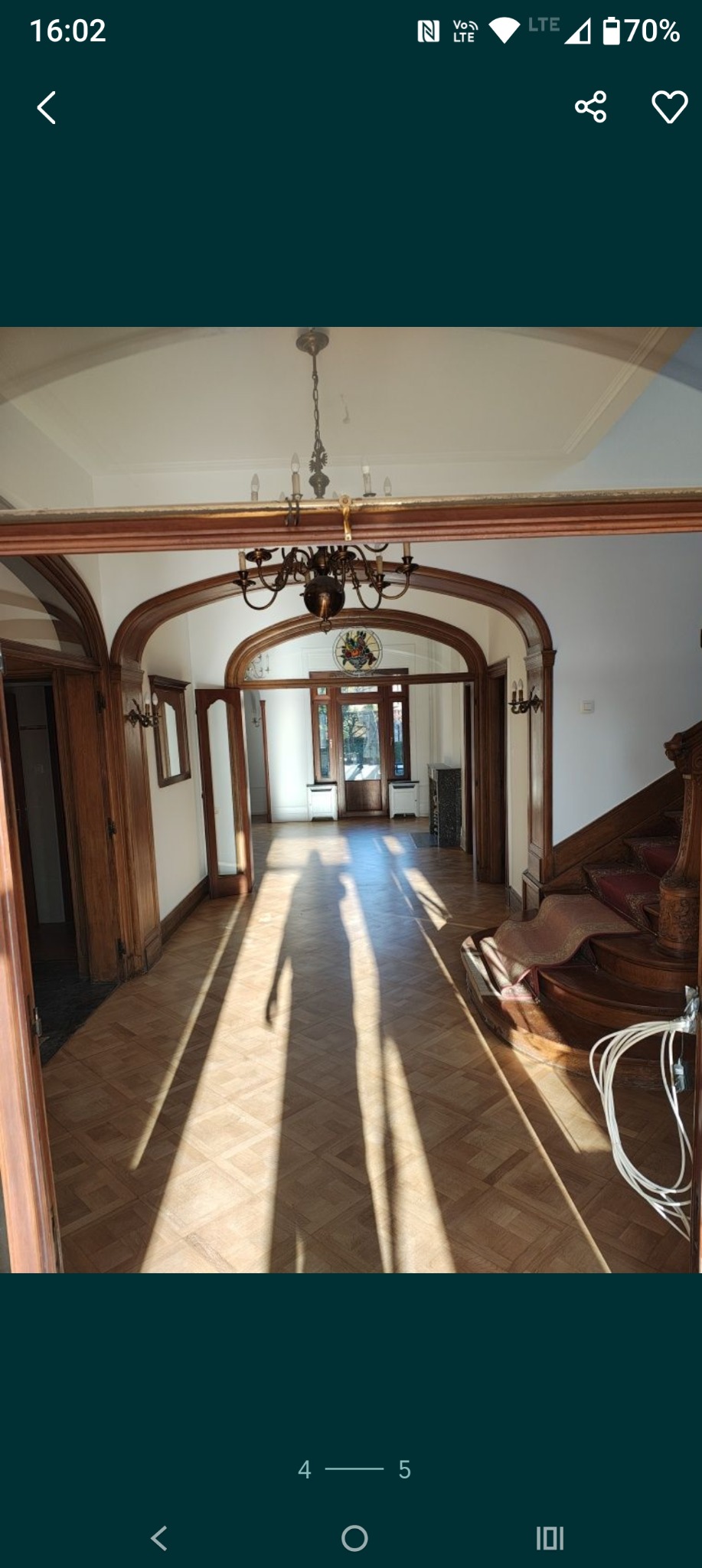 Jasne wnętrze domu z parkietem w jodełkę, drewnianą stolarką i schodami. Długie cienie na podłodze od popołudniowego słońca. Widoczne elementy dekoracyjne i stylowy żyrandol.