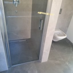 Katarzya Jarosław - Szklana kabina prysznicowa typu walk-in z odpływem liniowym w posadzce, wykończona szarymi płytkami imitującymi marmur, z widocznym fragmentem wiszącego bidetu i taśmą malarską zabezpieczającą szkło.