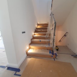 Katarzya Jarosław - Schody drewniane z podświetleniem LED w trakcie zabezpieczania przed pracami malarskimi, poręcz owinięta folią ochronną, widoczne taśmy malarskie i kartony na podłodze.