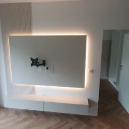 Nowoczesne wykończenie wnętrza: biała ściana z podświetlanym panelem TV, dekoracyjne lamele i szafka RTV, parkiet w jodełkę, otwarte drzwi do garderoby.