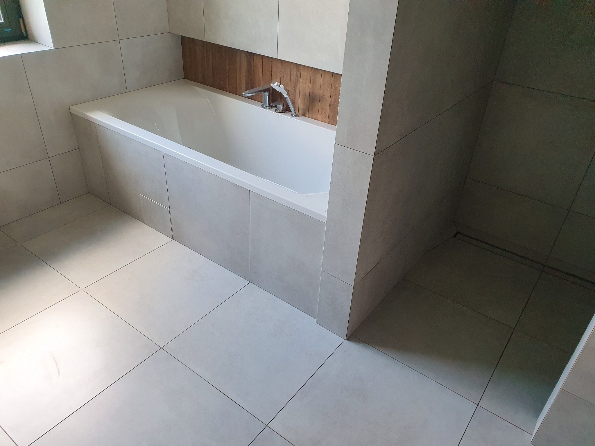 Wykończona łazienka z wanną obudowaną płytkami w odcieniach szarości, drewnopodobna wnęka nad wanną, chromowana bateria wannowa z słuchawką prysznicową, jasne płytki podłogowe i ścienne z fugami...