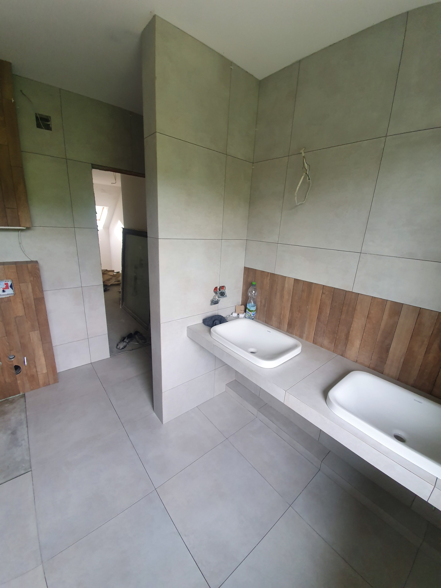 Łazienka w trakcie wykańczania: szare płytki na ścianach i podłodze, dwie białe umywalki na betonowym blacie, drewniana boazeria, widoczne przewody elektryczne, w tle otwarte drzwi do innego...