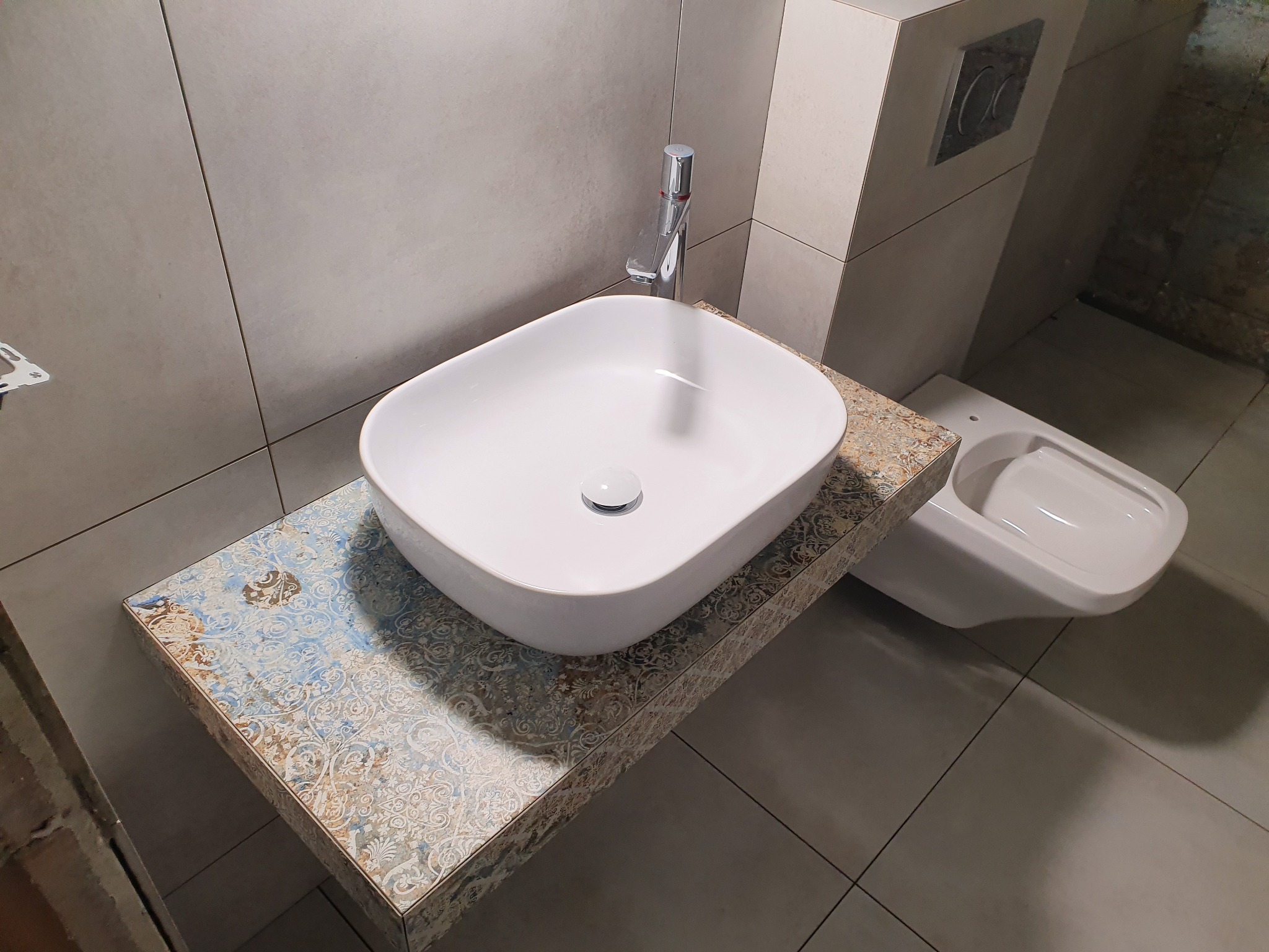 Nowoczesna, biała umywalka nablatowa na blacie z dekoracyjnym wzorem, chromowana bateria, wisząca toaleta w tle, szare płytki na ścianach i podłodze, minimalistyczny styl.