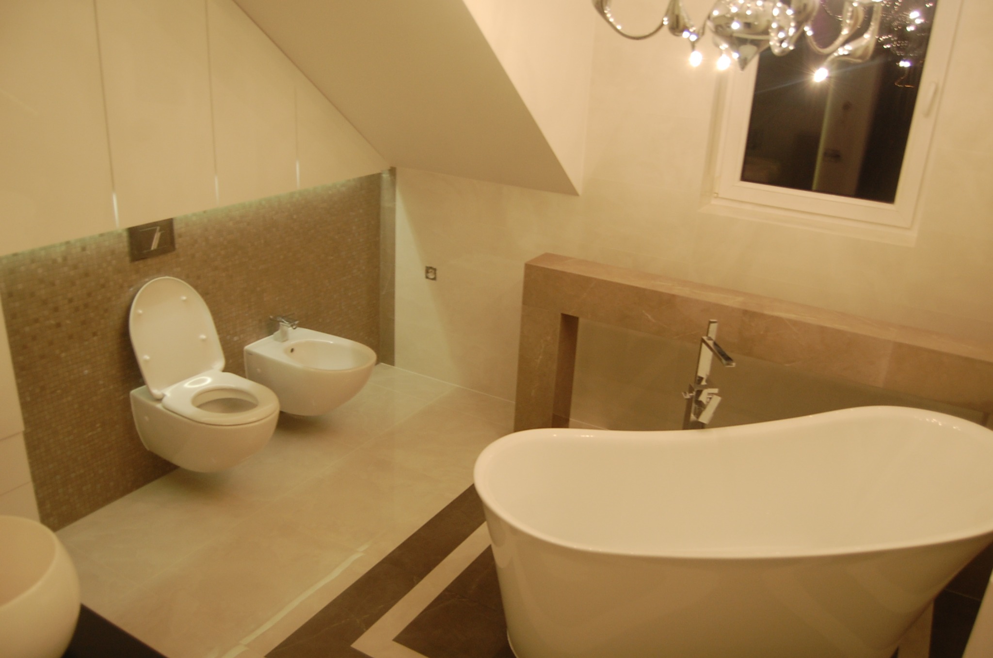 Nowoczesna łazienka na poddaszu z wolnostojącą wanną, wiszącą toaletą i bidetem na tle mozaiki ściennej, oświetlona żyrandolem z kryształkami.