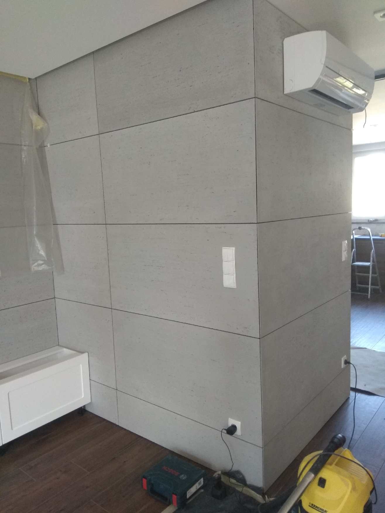 Wykończona łazienka z dużymi szarymi płytkami imitującymi kamień, biała szafka pod umywalkę, klimatyzator na ścianie, widoczne gniazdka elektryczne i narzędzia budowlane na podłodze.