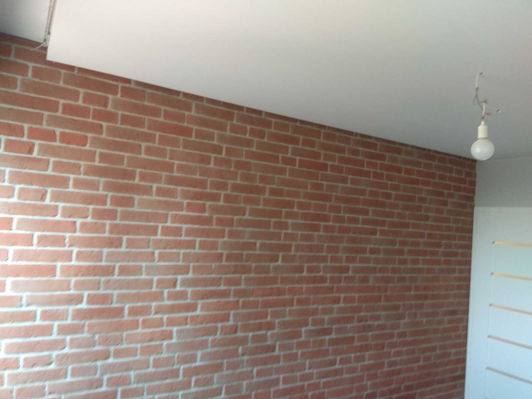 Ściana wykończona czerwoną cegłą w mieszkaniu w Krakowie, z widocznym białym sufitem i wiszącą żarówką. Ciekawa faktura i kolorystyka cegieł.