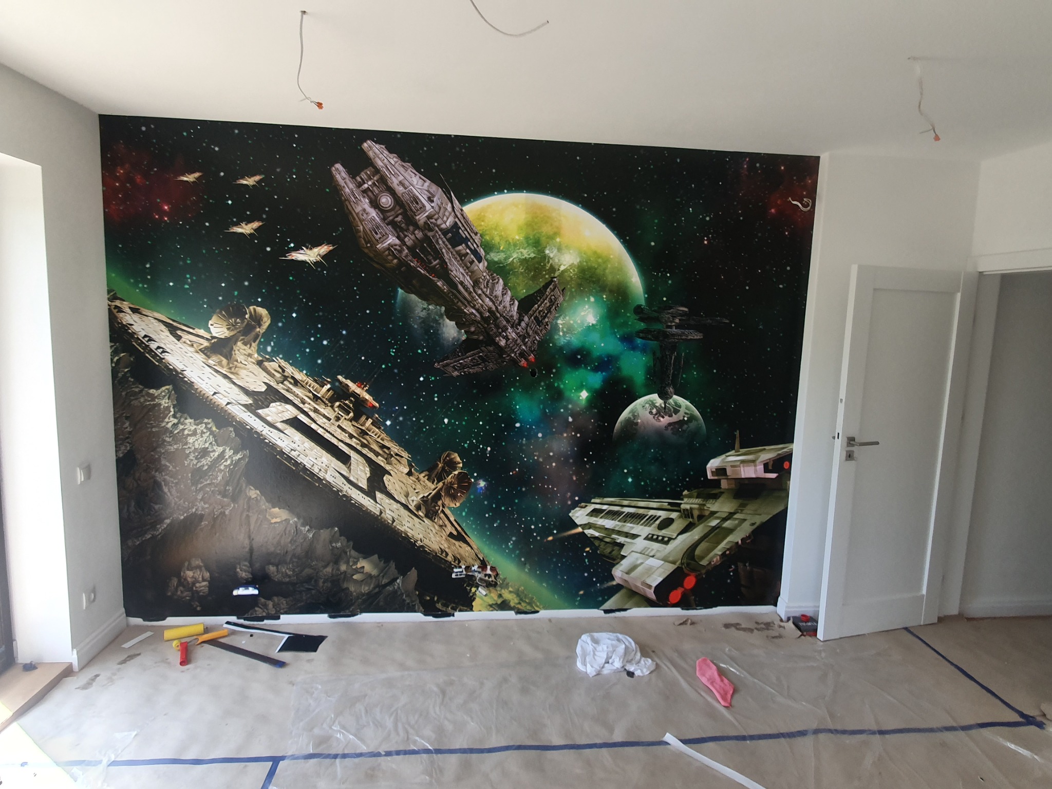 Pokój z fototapetą przedstawiającą kosmiczną bitwę, widoczne narzędzia i folia ochronna na podłodze sugerują prace wykończeniowe.