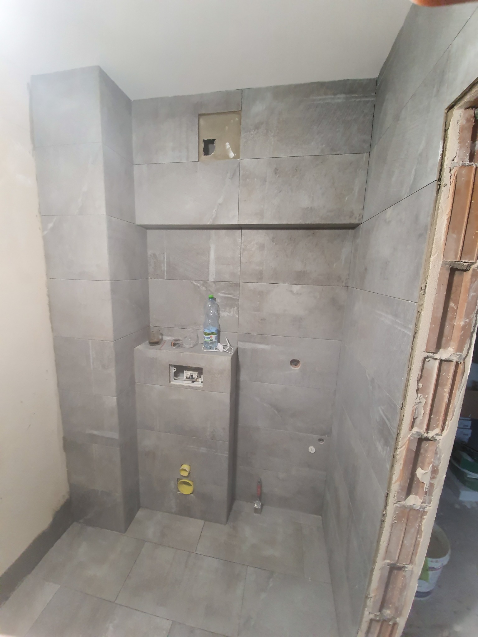 Łazienka w trakcie kładzenia szarych, prostokątnych płytek ceramicznych, widoczne niedokończone obramowanie drzwi i butelka wody na stelażu podtynkowym.