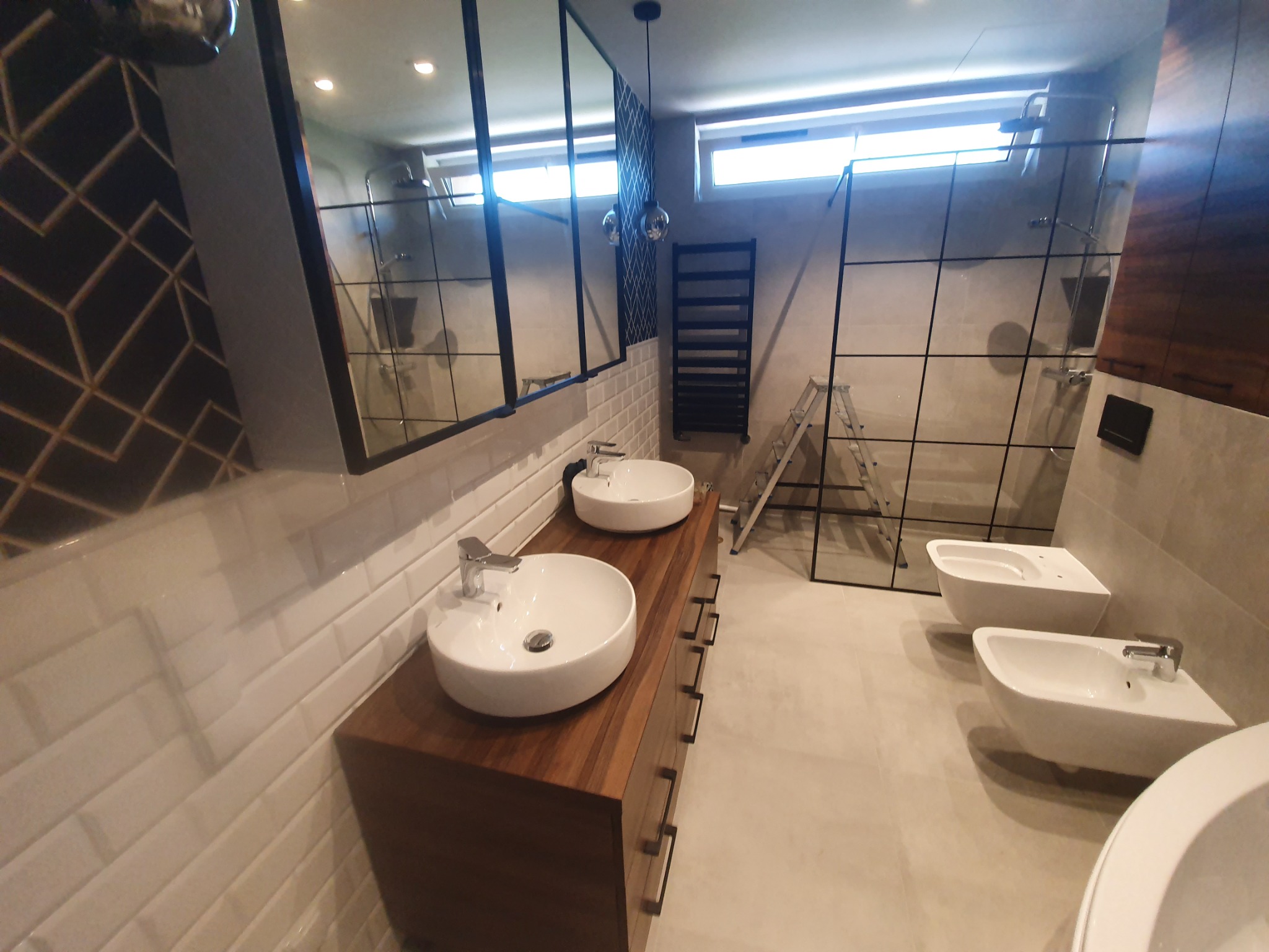 Nowoczesna łazienka z dwoma okrągłymi umywalkami na drewnianej szafce, białymi kaflami na ścianach, bidetami i kabiną prysznicową z czarnymi ramami, widoczna drabinka grzejnikowa i aluminiowa drabina.