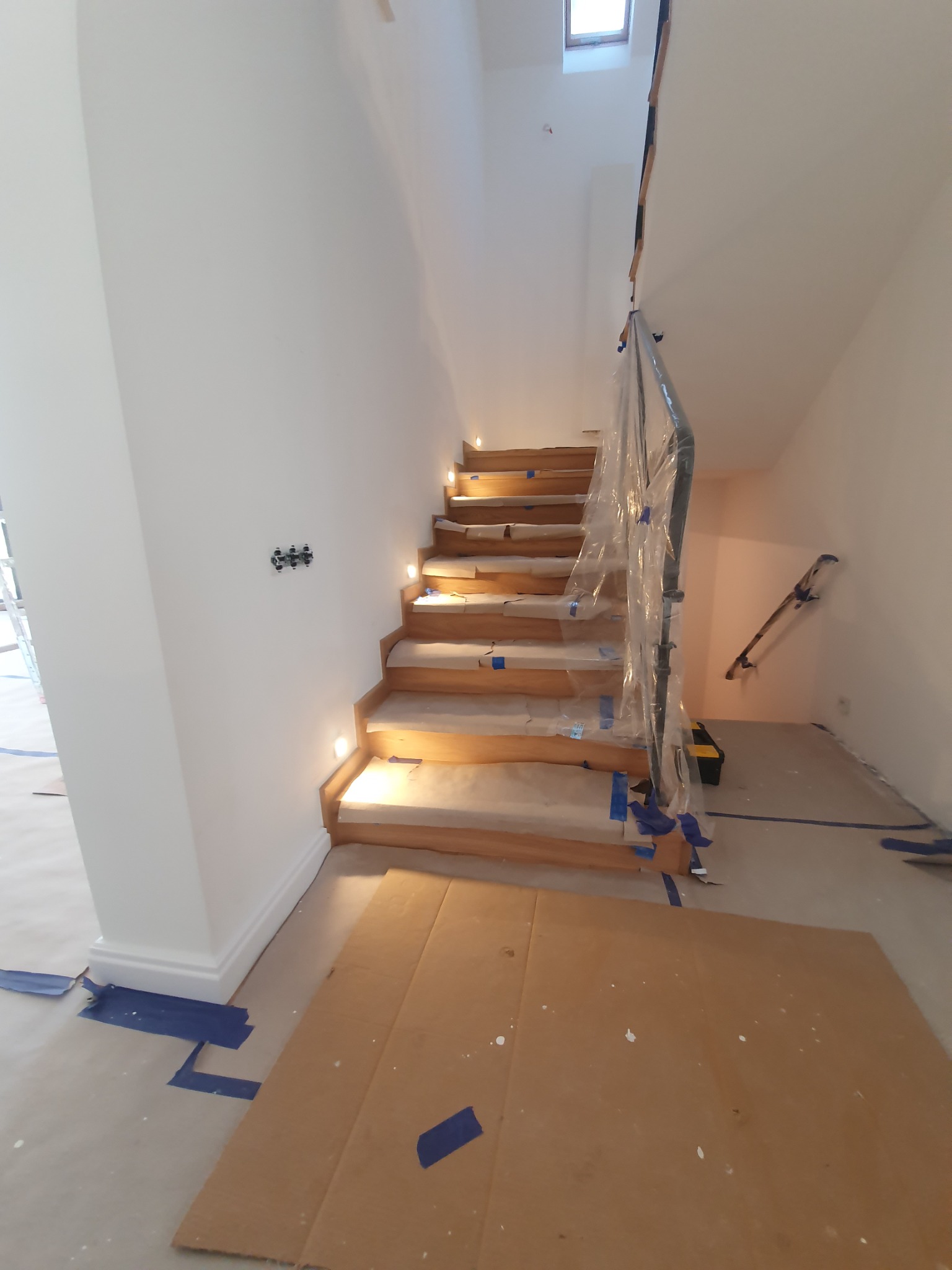 Schody drewniane z podświetleniem LED w trakcie zabezpieczania przed pracami malarskimi, poręcz owinięta folią ochronną, widoczne taśmy malarskie i kartony na podłodze.