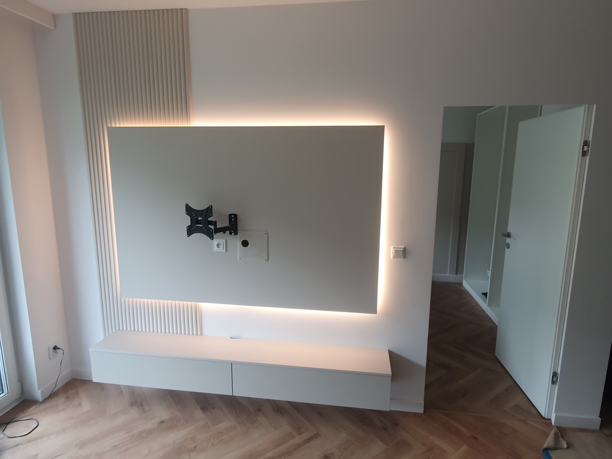 Nowoczesne wykończenie wnętrza: biała ściana z podświetlanym panelem TV, dekoracyjne lamele i szafka RTV, parkiet w jodełkę, otwarte drzwi do garderoby.