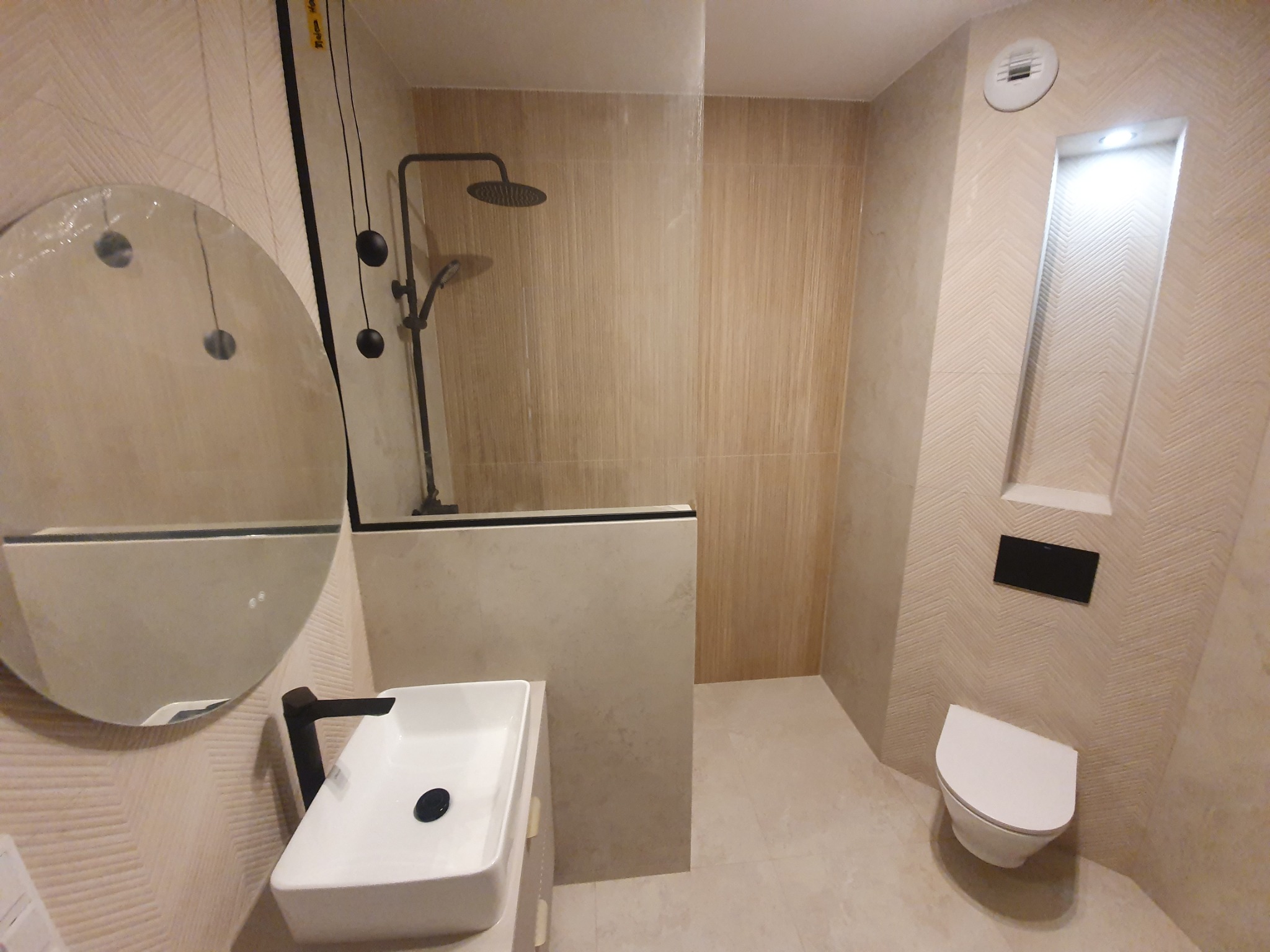 Nowoczesna łazienka z kabiną prysznicową typu walk-in, okrągłym lustrem, umywalką i podwieszaną toaletą. Beżowe płytki z subtelnym wzorem jodełki.