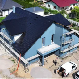 Niedokończony, dwukondygnacyjny budynek z ciemną dachówką, otoczony rusztowaniami; widoczne przygotowania do prac elewacyjnych, z hałdami piasku i żwiru na posesji.