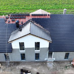 LEXO SP. Z O.O. - Widok z góry na budowę domu z czarną dachówką, częściowo ułożoną, z pracownikiem na dachu, widoczne rusztowanie, niezadaszona część domu, oraz przenośna toaleta na działce.