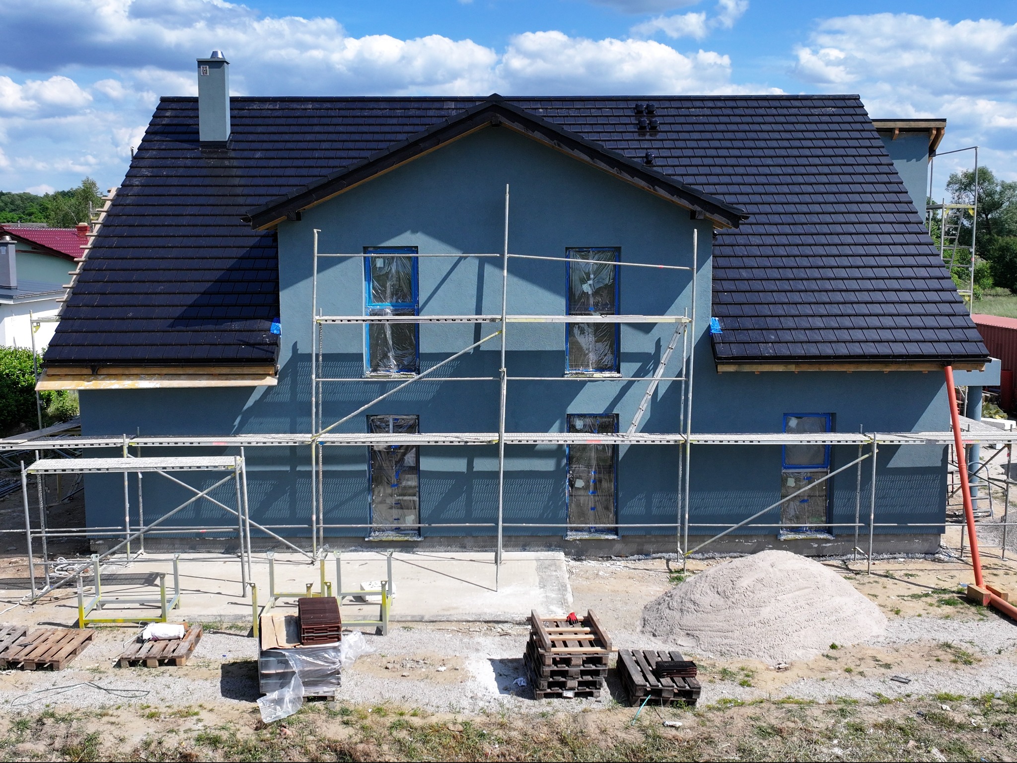 Dwukondygnacyjny budynek w trakcie prac wykończeniowych elewacji, z widocznym rusztowaniem, zabezpieczonymi oknami folią, stertą piasku i materiałami budowlanymi na paletach.