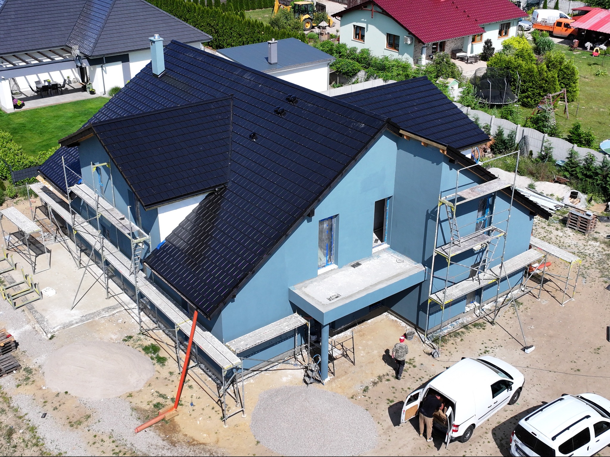 Niedokończony, dwukondygnacyjny budynek z ciemną dachówką, otoczony rusztowaniami; widoczne przygotowania do prac elewacyjnych, z hałdami piasku i żwiru na posesji.