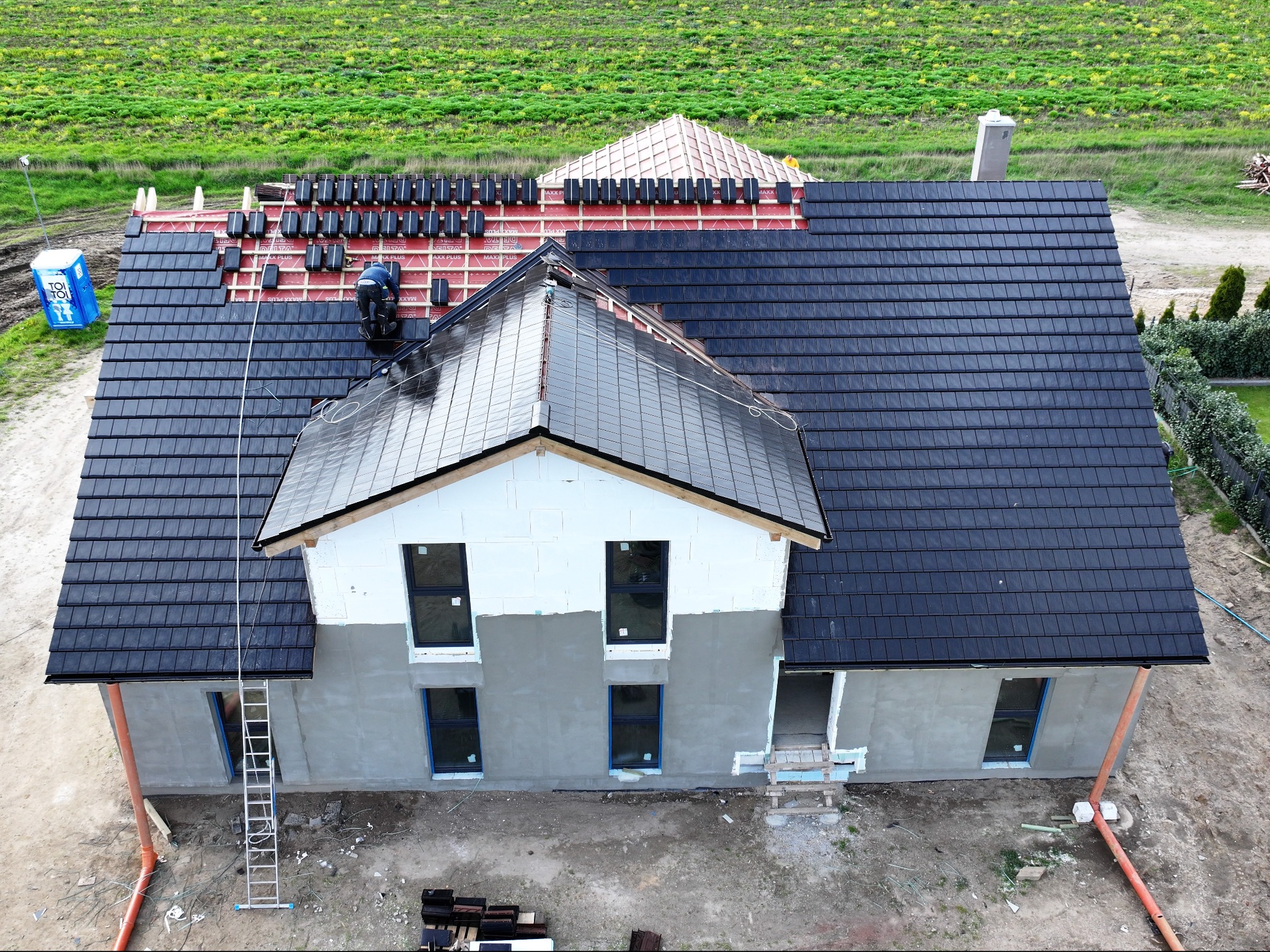 Widok z góry na budowę domu z czarną dachówką, częściowo ułożoną, z pracownikiem na dachu, widoczne rusztowanie, niezadaszona część domu, oraz przenośna toaleta na działce.