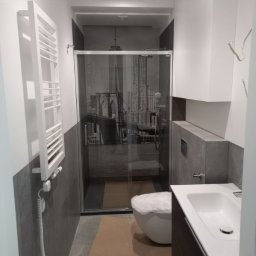 Viktor Yevfymets - Nowoczesna łazienka w szarościach z kabiną prysznicową z motywem nowojorskiego mostu Brooklyńskiego, biała umywalka z szafką, grzejnik drabinkowy oraz zabezpieczona folią toaleta.