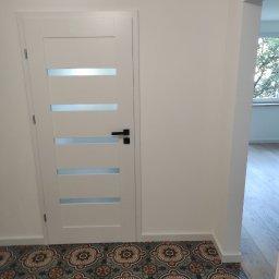 Viktor Yevfymets - Białe drzwi wewnętrzne z poziomymi przeszkleniami na tle wzorzystej, ceramicznej podłogi w odcieniach brązu i zieleni, widok na sąsiednie pomieszczenie z drewnianą podłogą.