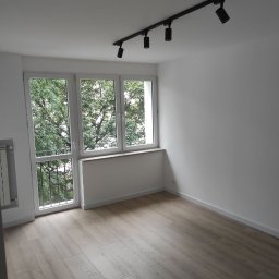 Viktor Yevfymets - Pusty, odnowiony pokój z panelami podłogowymi w odcieniach szarości, białymi ścianami, oknem z widokiem na drzewa i czarną szyną oświetleniową z czterema reflektorami na suficie.