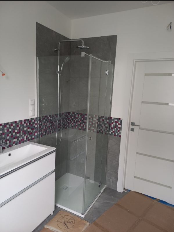 Nowoczesna łazienka po remoncie: kabina prysznicowa ze szklanymi drzwiami, biała umywalka z szafką, szare płytki imitujące kamień i dekoracyjny pas mozaiki w odcieniach szarości, bieli i fioletu.