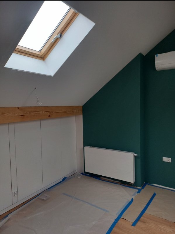 Poddasze po malowaniu na butelkową zieleń, zabezpieczona folią podłoga, białe szafki w zabudowie i grzejnik, okno dachowe w drewnianej ramie.