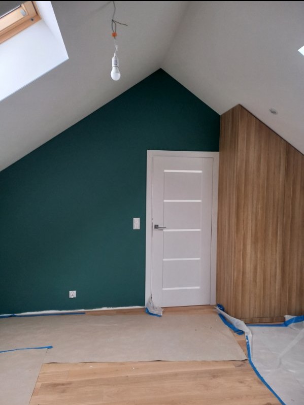Pokój na poddaszu w trakcie malowania ścian na ciemny zielony kolor, zabezpieczona podłoga, białe drzwi z czterema poziomymi panelami, zabudowa z paneli drewnopodobnych, surowa żarówka wisząca...