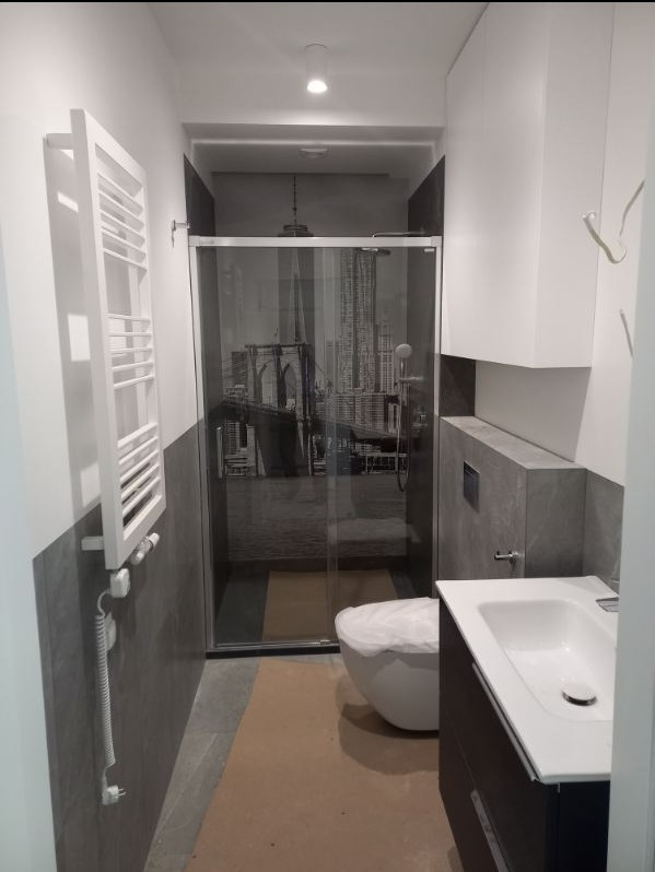 Nowoczesna łazienka w szarościach z kabiną prysznicową z motywem nowojorskiego mostu Brooklyńskiego, biała umywalka z szafką, grzejnik drabinkowy oraz zabezpieczona folią toaleta.
