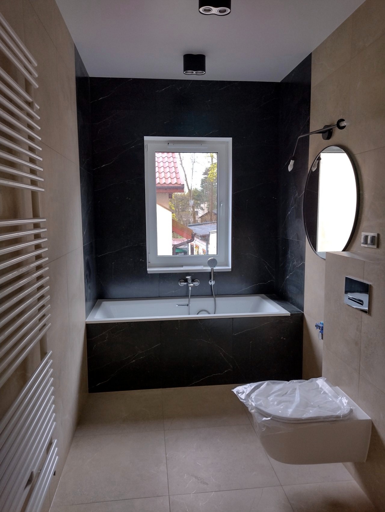 Nowoczesna łazienka w beżowo-czarnej kolorystyce z wanną obudowaną czarnym marmurem, grzejnikiem drabinkowym i podwieszaną toaletą w folii ochronnej, widok na dom przez okno.