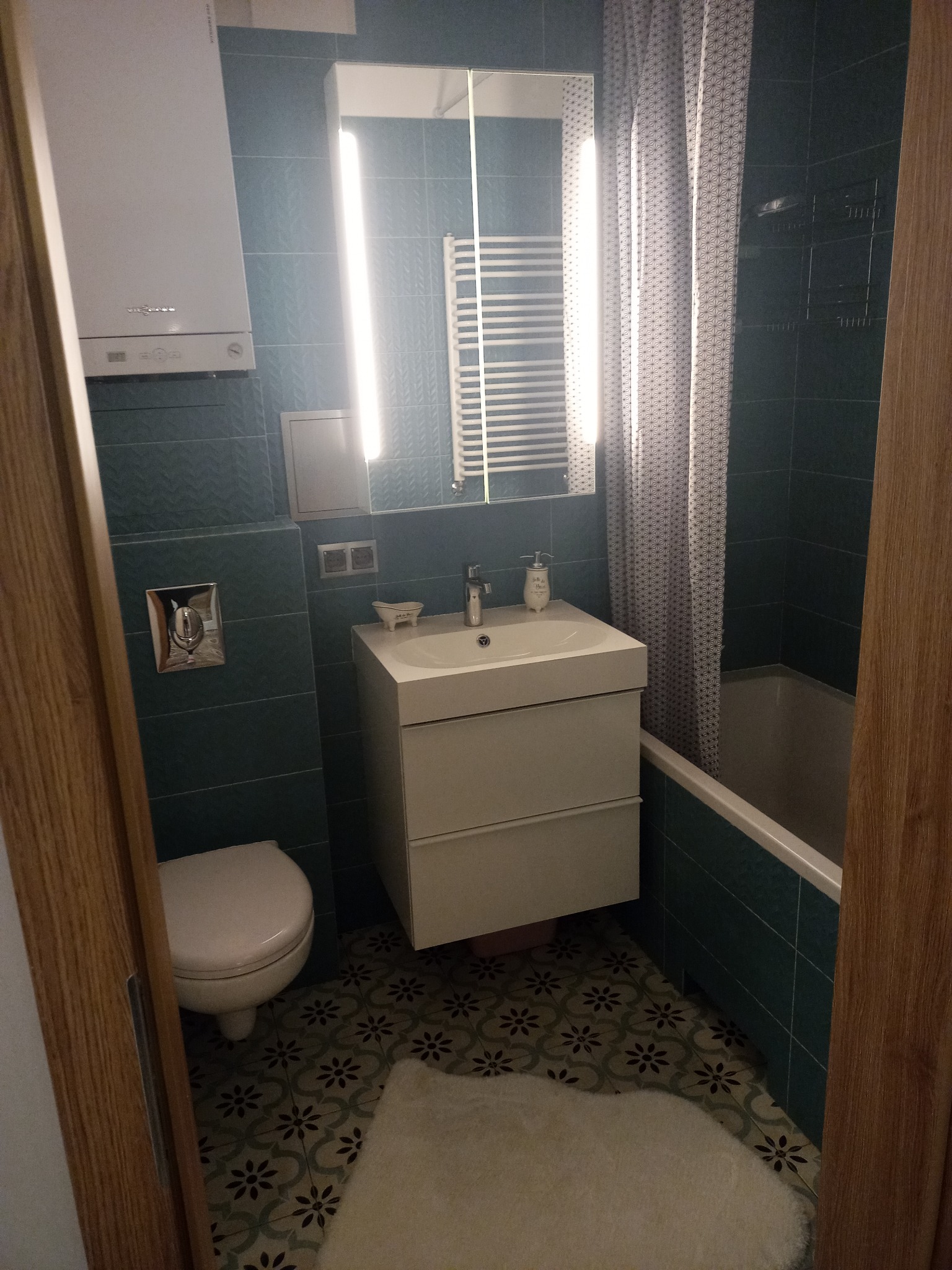 Nowoczesna łazienka z turkusowymi płytkami, białym blatem umywalki z szafką, wiszącą toaletą i grzejnikiem drabinkowym, oświetlona listwą LED nad lustrem.