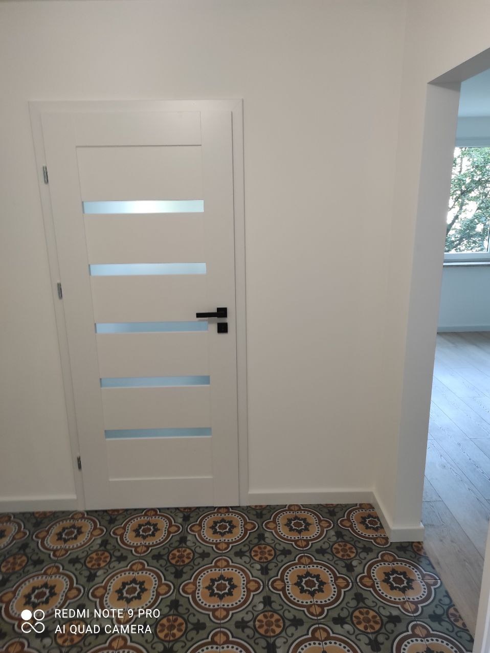 Białe drzwi wewnętrzne z poziomymi przeszkleniami na tle wzorzystej, ceramicznej podłogi w odcieniach brązu i zieleni, widok na sąsiednie pomieszczenie z drewnianą podłogą.