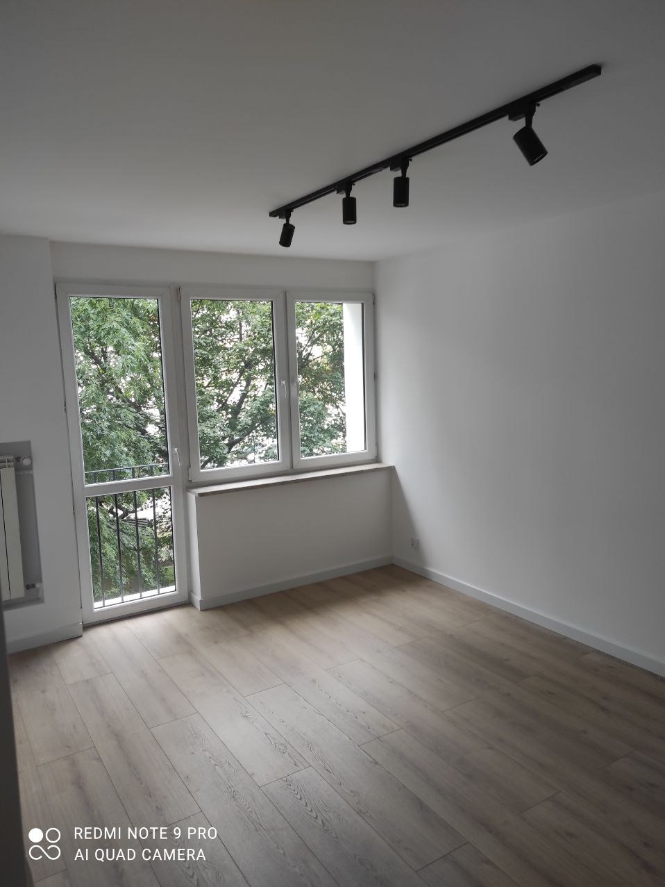 Pusty, odnowiony pokój z panelami podłogowymi w odcieniach szarości, białymi ścianami, oknem z widokiem na drzewa i czarną szyną oświetleniową z czterema reflektorami na suficie.