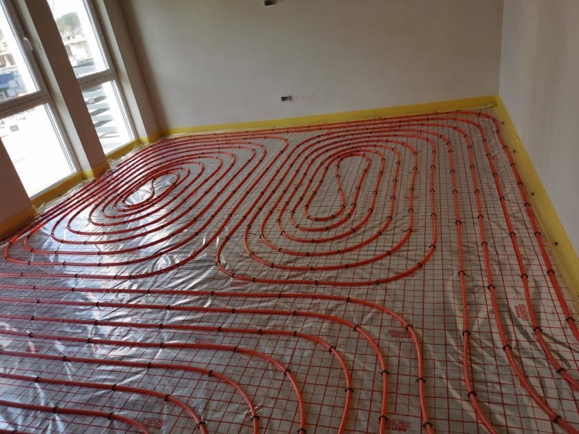 Pomarańczowe rury ogrzewania podłogowego ułożone spiralnie na siatce, zabezpieczone folią, widok z góry na niedokończoną instalację w jasnym pomieszczeniu z oknem.