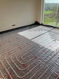 Rozłożone rury ogrzewania podłogowego na folii izolacyjnej, widoczne mocowania i fragment ściany z zaznaczonym miejscem na instalację.