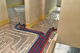 Rozłożone rury ogrzewania podłogowego na folii izolacyjnej w trakcie montażu. Widoczne niebieskie i czerwone rury, białe uchwyty mocujące oraz fragmenty ścian w surowym stanie.