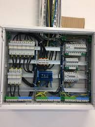 Otwarta skrzynka elektryczna z widocznymi bezpiecznikami, przewodami i listwami zaciskowymi, zamontowana na białej ścianie, z rurami instalacyjnymi w tle.